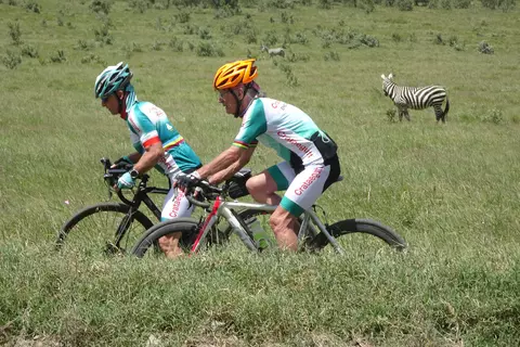 Zwei Radfahrer auf Mountainbikes mit einem Zebra im Hintergrund.