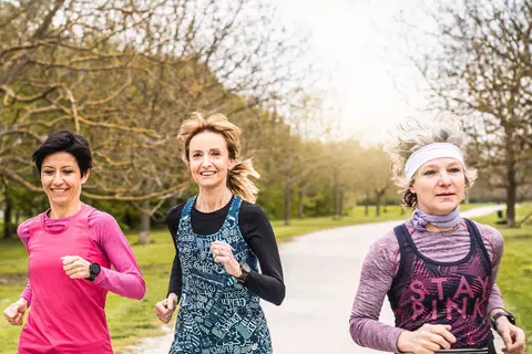 Drei Frauen mittleren Alters joggen zusammen auf einem Weg in einem Park, der von Bäumen umgeben ist.