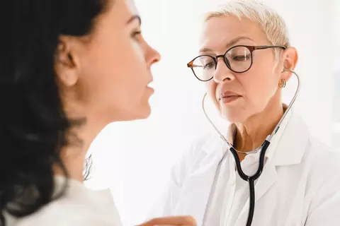 Ärztin mit Kurzhaarschnitt und Brille verwendet ein Stethoskop, um eine Patientin während einer medizinischen Untersuchung abzuhören.